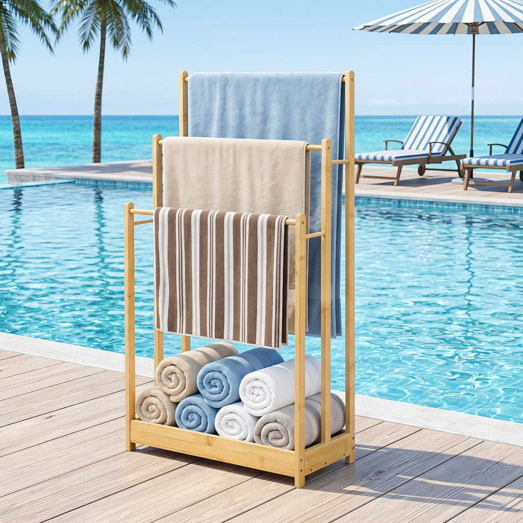 A bamboo towel rack set on a wooden deck beside a swimming pool, holding three towels and several rolled towels underneath. A beach poolside scene with lounge chairs, palm trees, and ocean water is visible in the background.