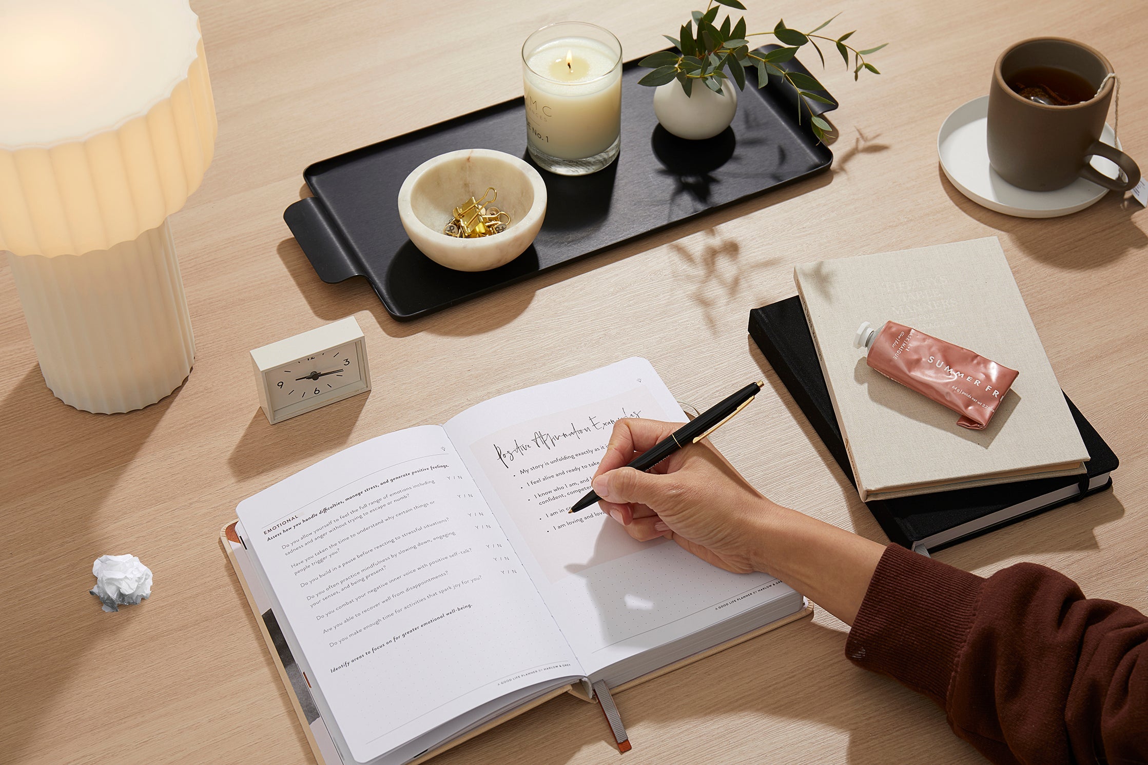 Person writing in a notebook on a wooden desk with a candle, plant, and coffee cup in the background.