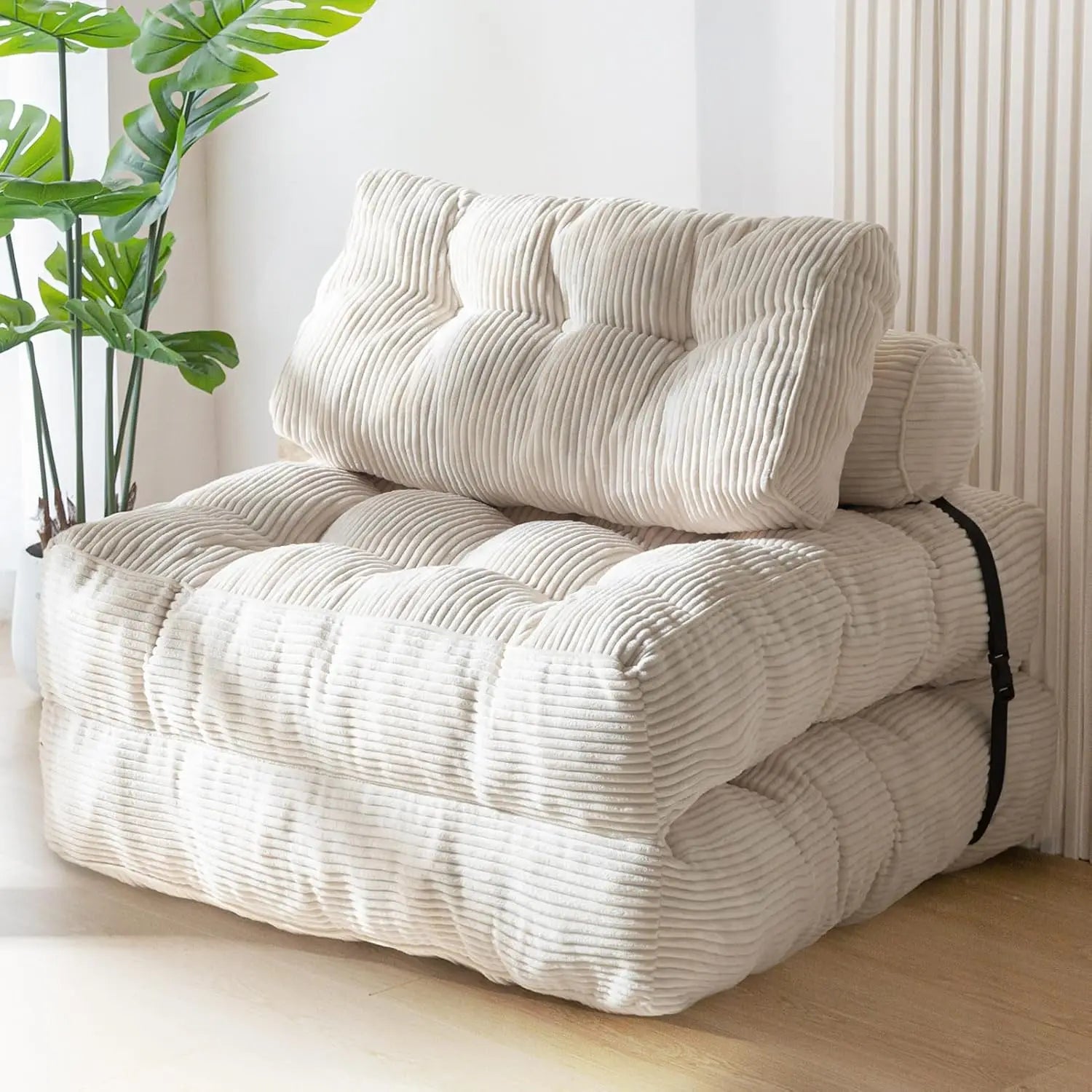 A cream-colored corduroy folding sofa bed with a matching back cushion, styled in a bright room with a wooden floor, a vertical slatted wall panel, and a potted green plant.