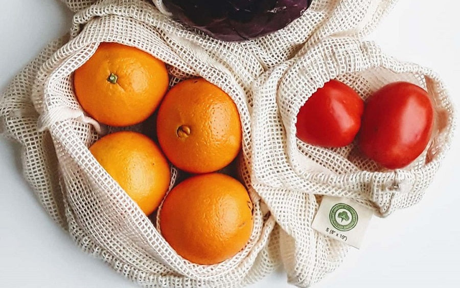 Two reusable mesh produce bags filled with fresh fruit and vegetables on a white background. One bag contains three oranges, and the other contains two red tomatoes.